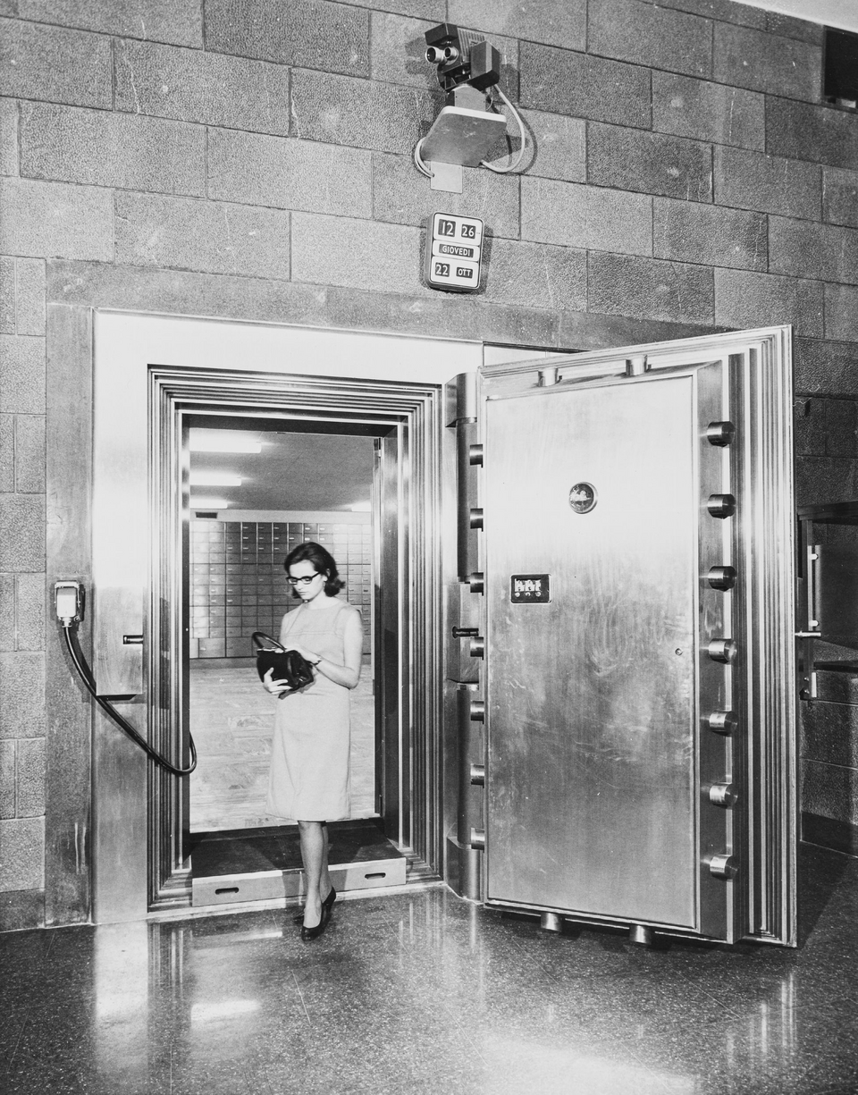 A steel bank vault door standing open, with rows of safe deposit boxes in the room beyond. Photographed in 1967.